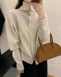 Woman buttons latte 100 % cashmere high-neck cardigan on Paris street, ribbed placket and tortoise buttons give effortless French polish.