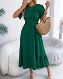 Model wearing ruffle trim short sleeve midi dress with shirred waist and tiered hem, carrying woven straw handbag, white strappy sandals, indoor cozy setting.
