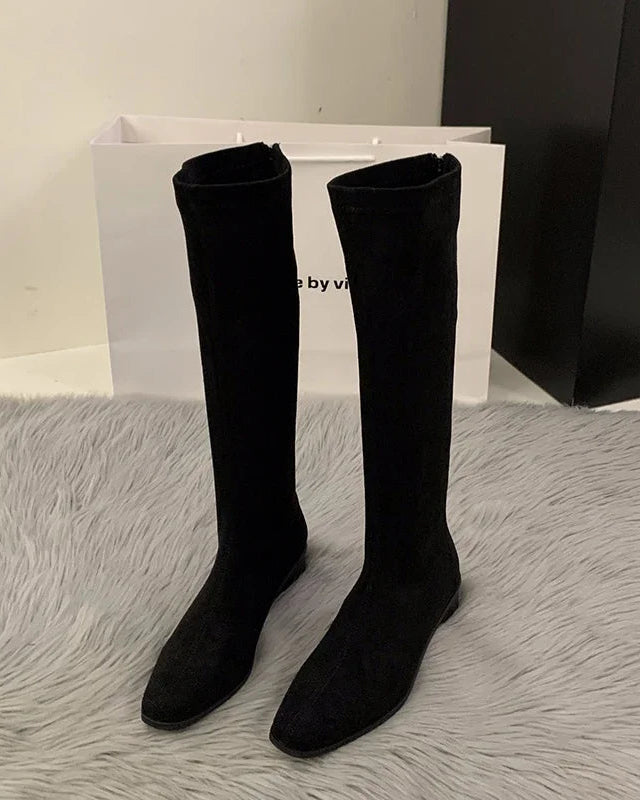 A pair of vintage stretch knee-high boots featuring a sleek flock upper, a modern square toe, and a low block heel for stable, all-day comfort.