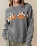 A pumpkin bow knit pullover with pumpkin and bow motifs, loose fit and long sleeves, designed for a cozy winter layering look that pairs from sneakers to boots.