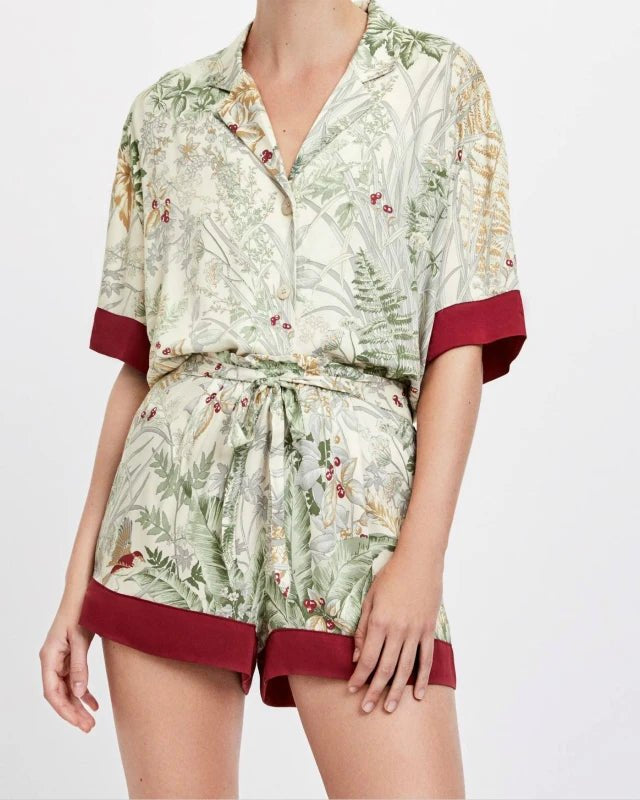 Viscose Satin pajama set featuring a tropical print with birds, ferns, and red berries. This two-piece set includes a short-sleeve, button-down shirt with a turn-down collar and contrasting burgundy trim, paired with matching shorts that have a drawstring waist.